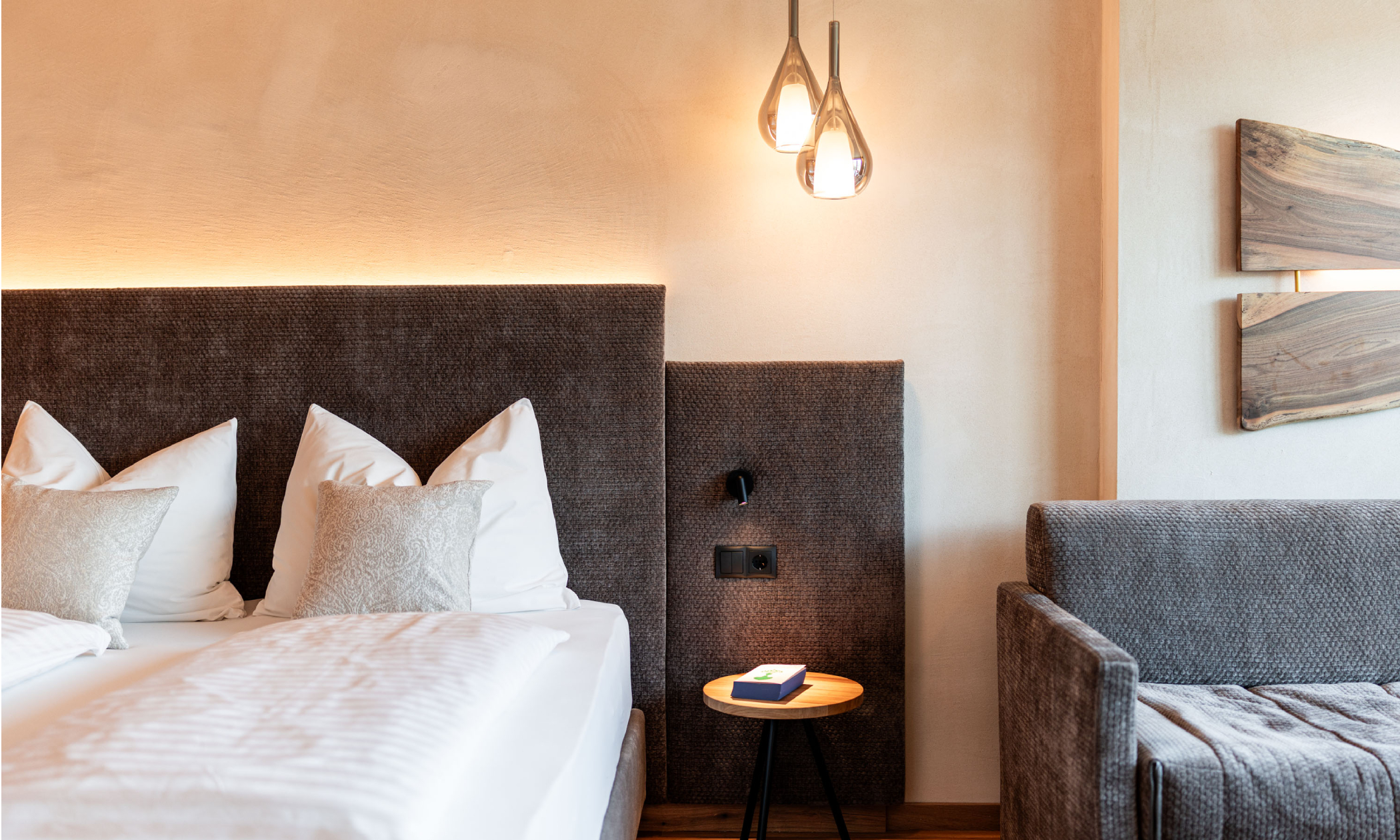 ein sauber gemachtes Bett, eine braune Couch, ein Buch auf dem kleinen Tisch neben dem Bett - Egger Apartments & Suites