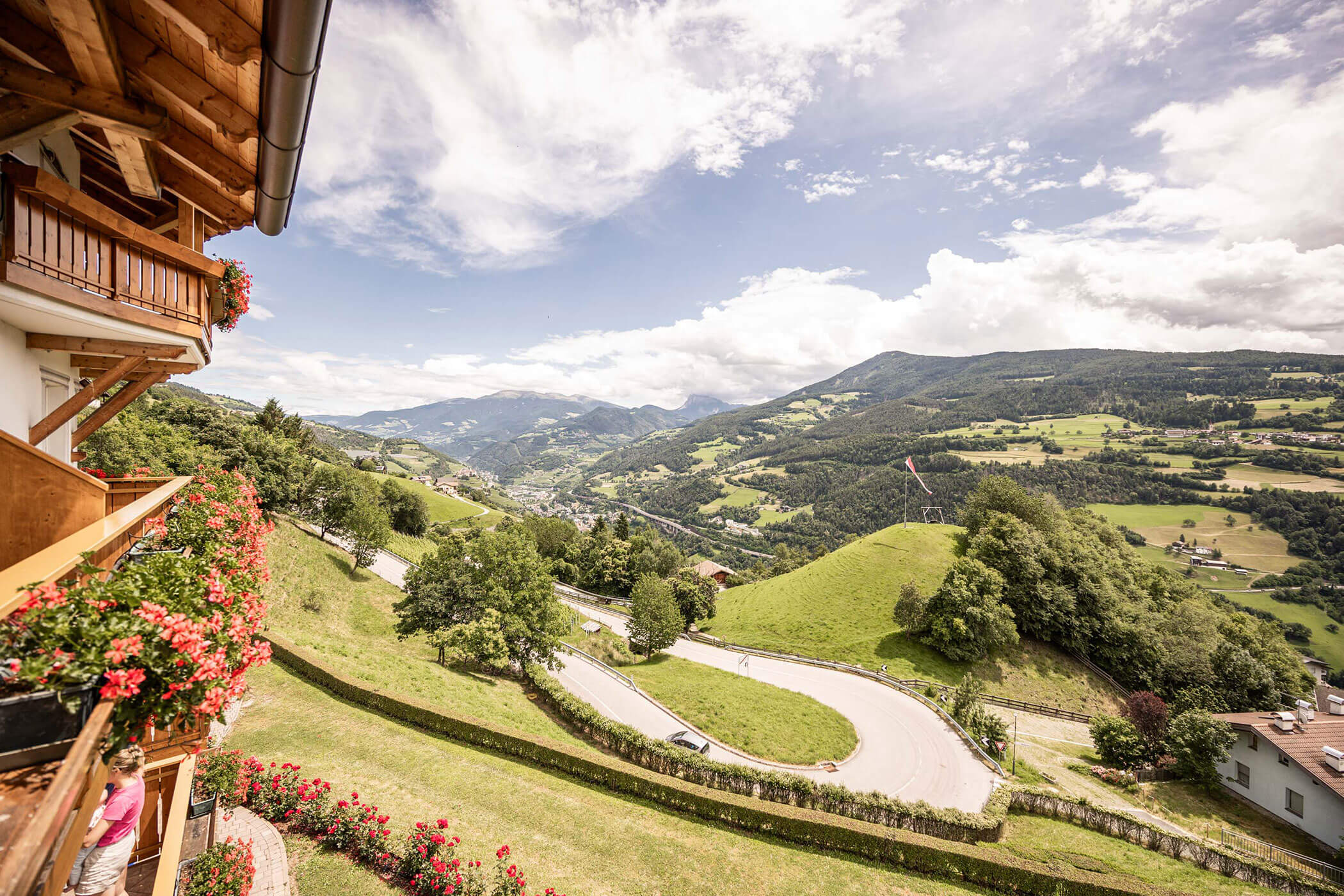 Der Balkon mit roten Blumen bietet einen Blick auf eine kurvenreiche Straße, grüne Hügel und ferne Berge unter einem teilweise bewölkten Himmel. - Egger Apartments & Suites