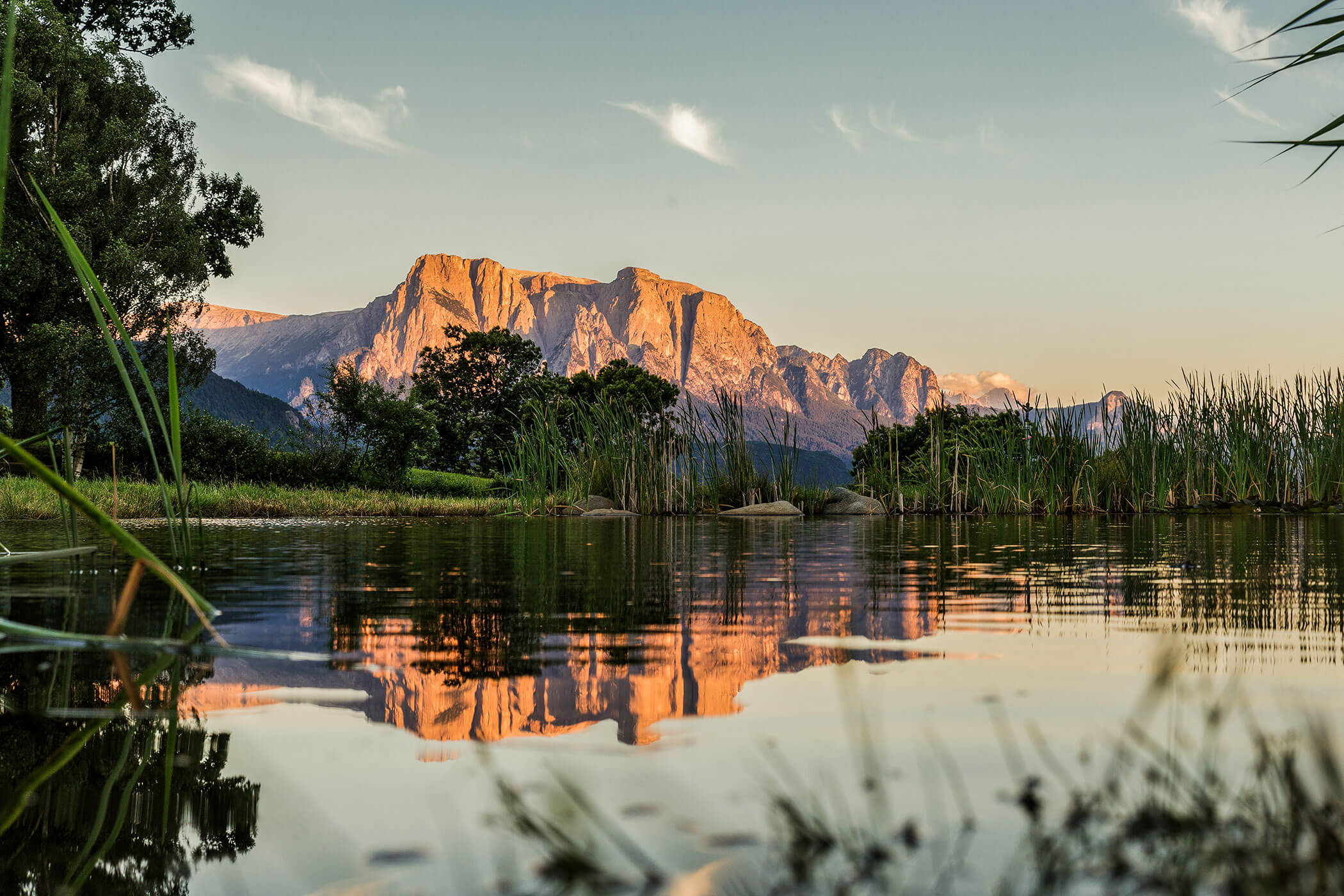 Ein Berg bei Sonnenuntergang, der sich in einem ruhigen Teich spiegelt, umgeben von Schilf und grünen Bäumen unter einem klaren Himmel. - Egger Apartments & Suites