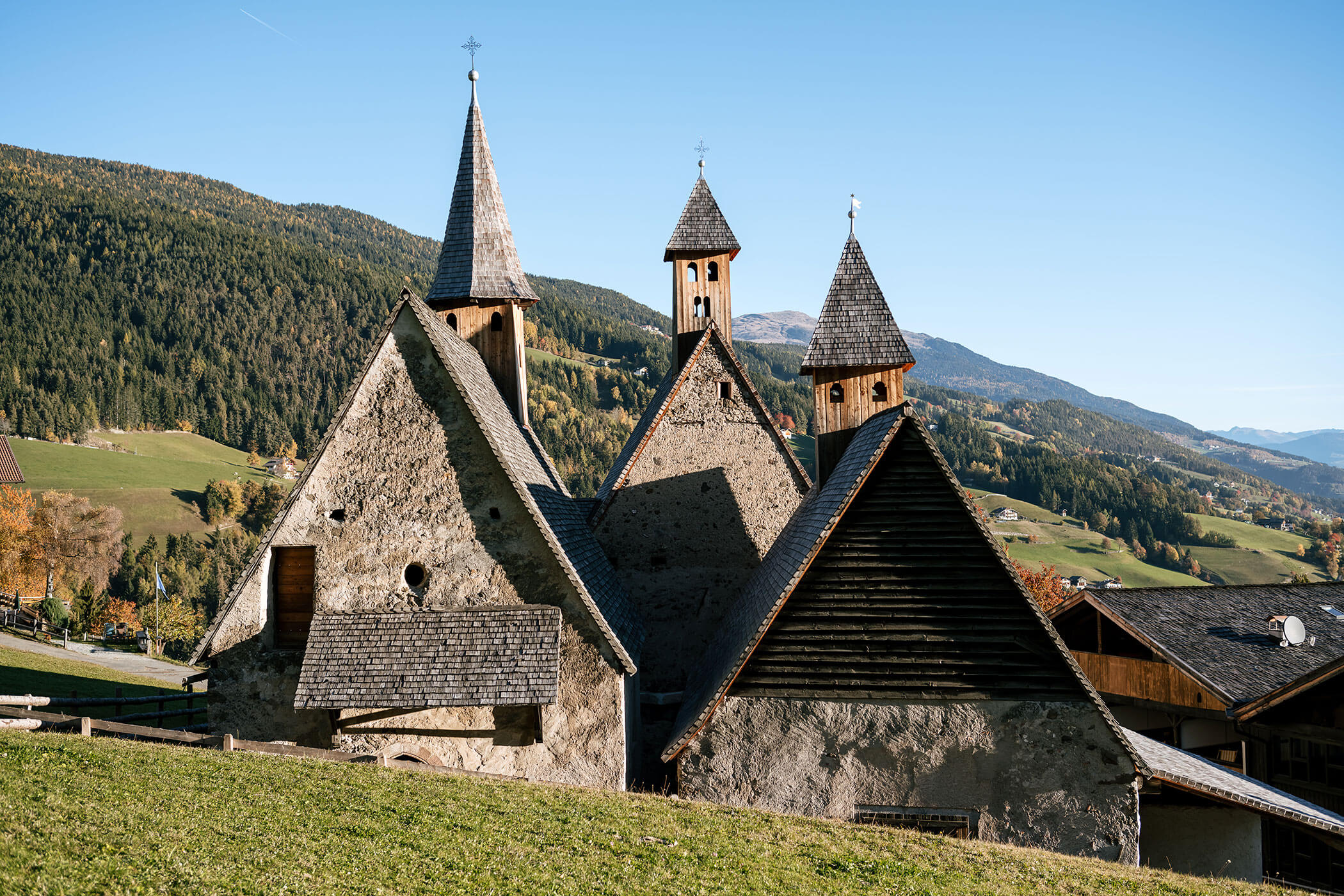 Alte Steinkirche mit drei spitzen Türmen auf einer grasbewachsenen Anhöhe, umgeben von Bergen und Bäumen. - Egger Apartments & Suites