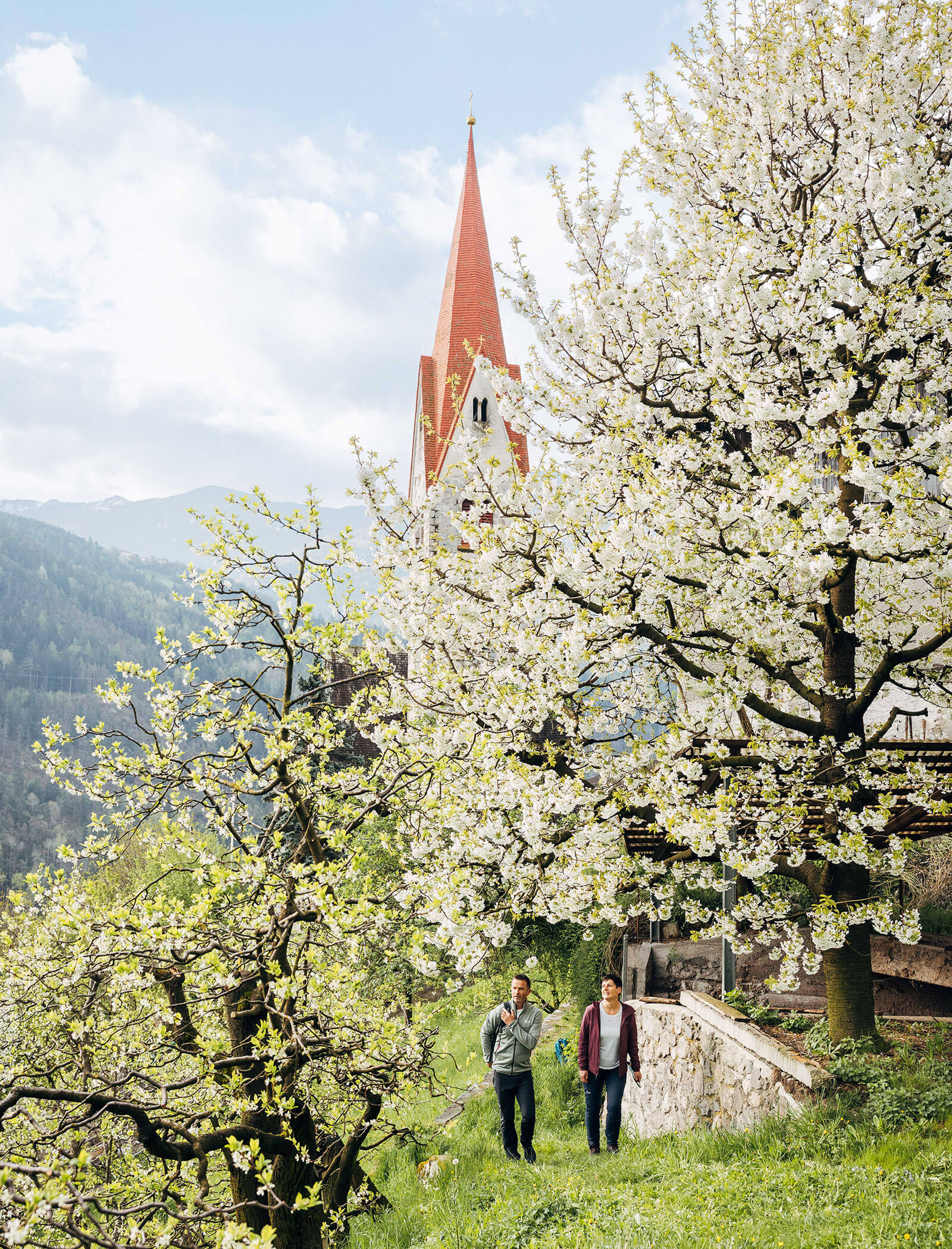 Zwei Personen gehen an blühenden Bäumen in der Nähe einer Kirche mit einem hohen roten Kirchturm in einer bergigen Landschaft vorbei. - Egger Apartments & Suites