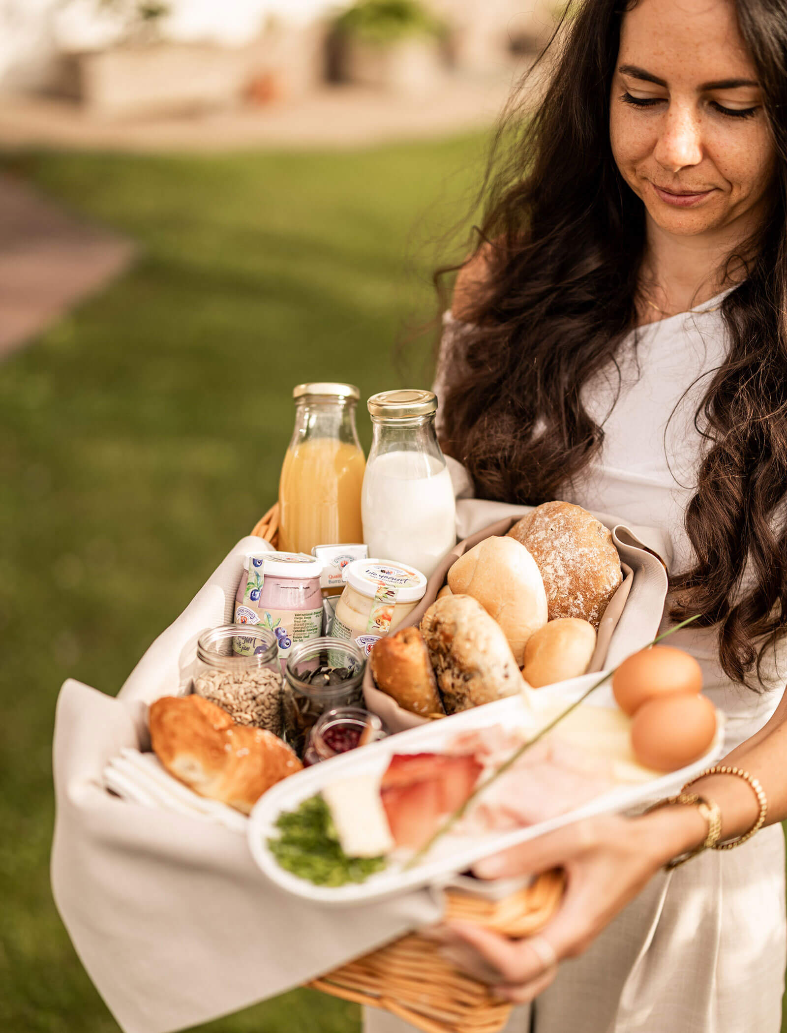 Eine Frau trägt einen Korb mit Brot, Eiern, Saft, Milch und verschiedenen Frühstücksprodukten ins Freie. - Egger Apartments & Suites