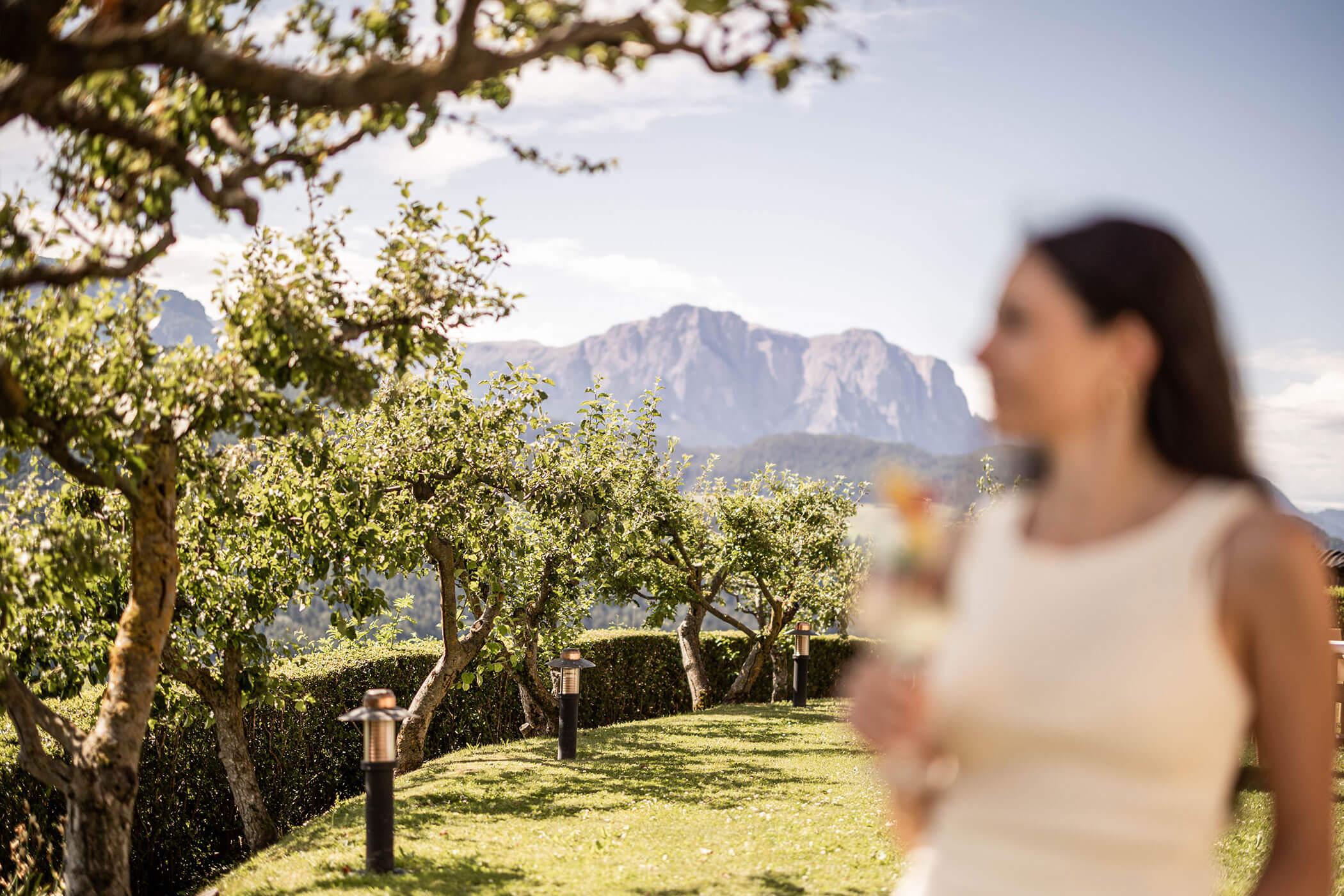 Eine Frau mit einem Getränk in der Hand steht in einem sonnigen Obstgarten, im Hintergrund sind die Berge zu sehen. - Egger Apartments & Suites