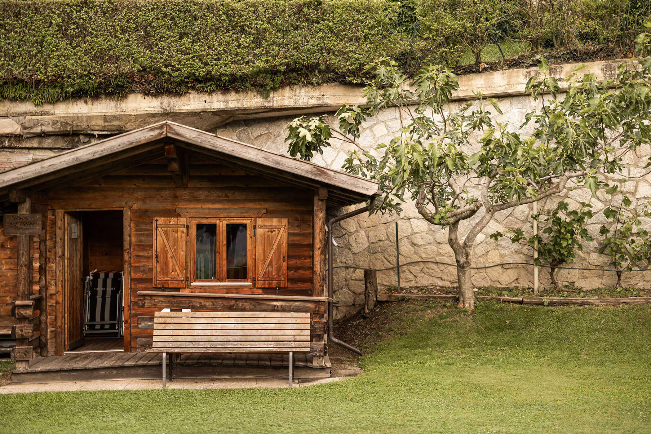 Eine kleine Holzhütte mit offenen Fensterläden, einer Bank und einem Baum davor, eingefasst von einer Steinmauer und Gras. - Egger Apartments & Suites