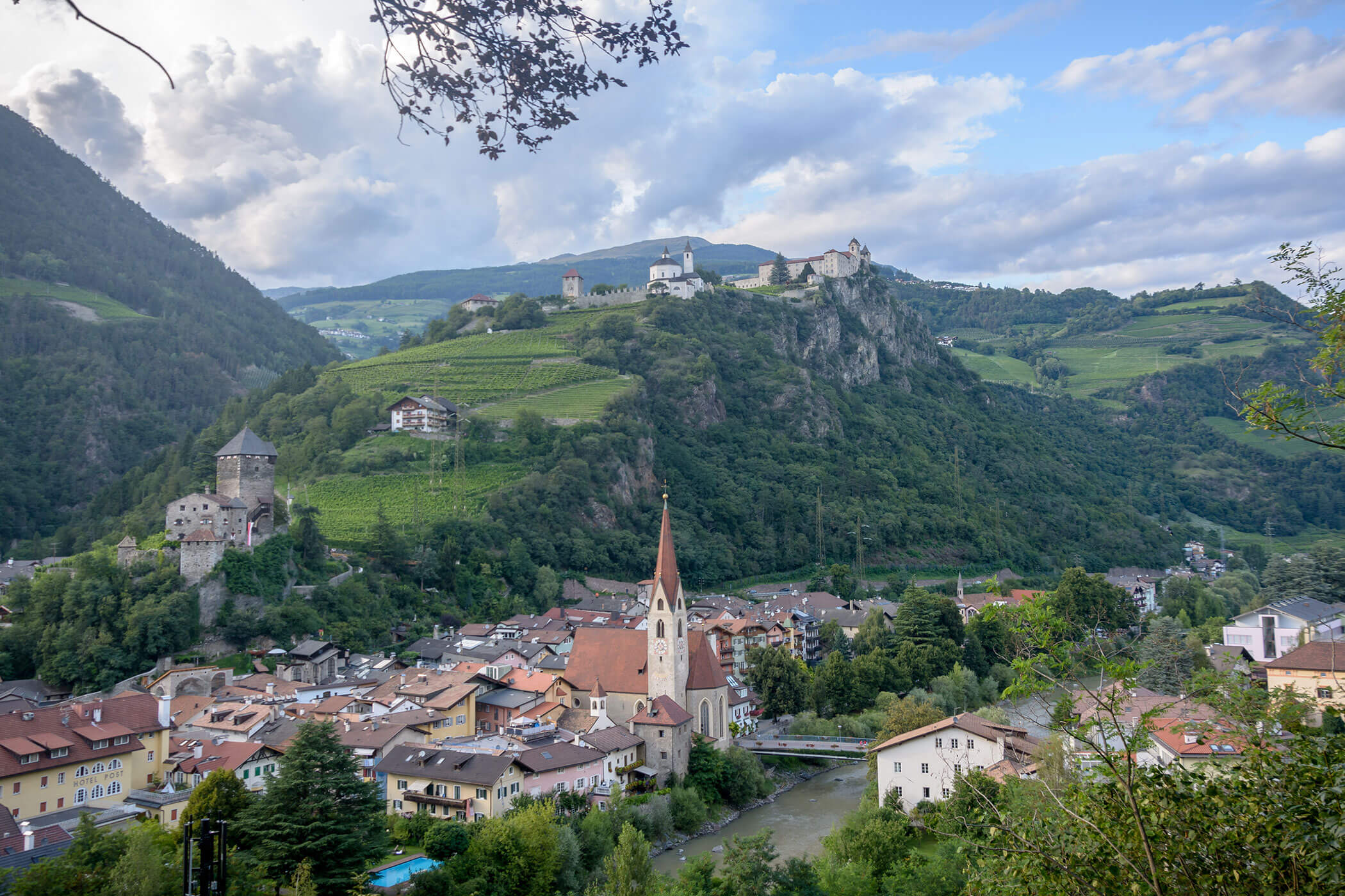 Ein Dorf mit einer Kirche, umgeben von Hügeln, Schlössern und Grünflächen unter einem teilweise bewölkten Himmel. - Egger Apartments & Suites