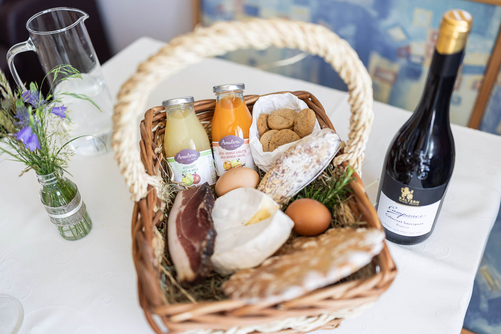 Ein Weidenkorb mit Saft, Eiern, Brot, Käse, Keksen und Wurstwaren auf einer weißen Tischdecke. - Egger Apartments & Suites