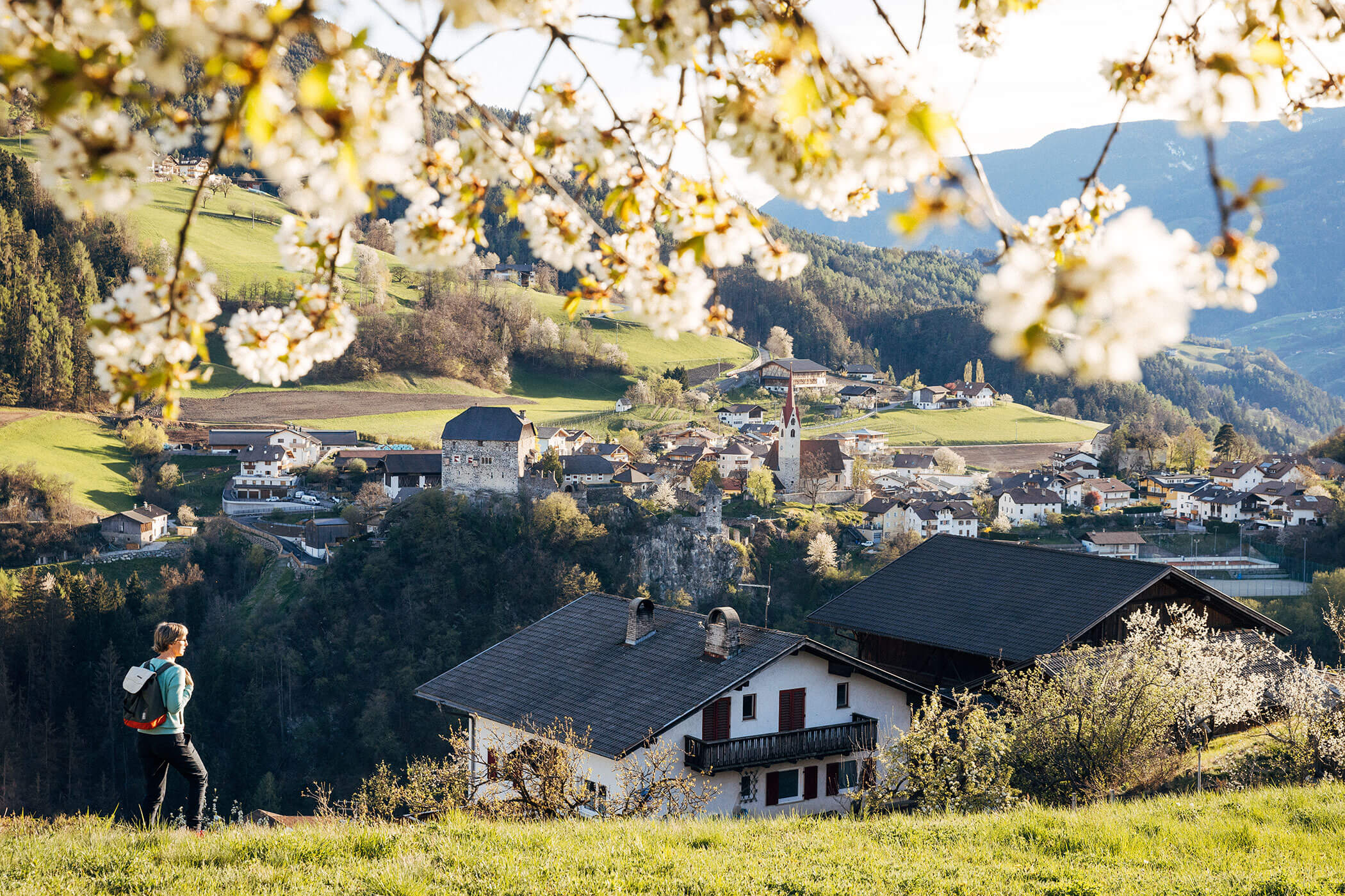 Eine Person mit Rucksack geht in der Nähe eines Hauses mit Blick auf ein Dorf und die Berge, umrahmt von blühenden Ästen. - Egger Apartments & Suites