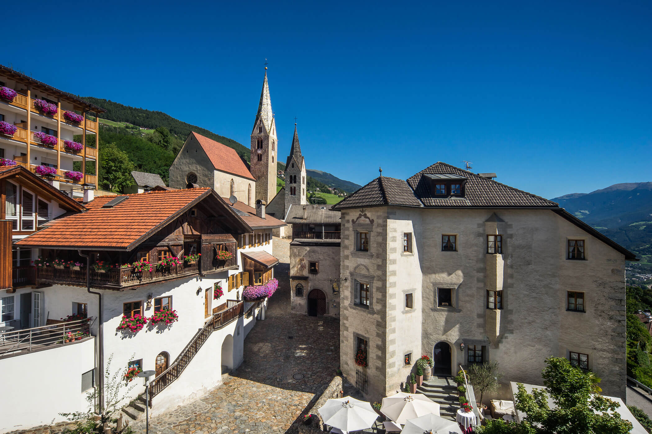 Ein malerischer Dorfplatz mit historischen Gebäuden, bunten Blumen und zwei hohen Kirchtürmen unter einem blauen Himmel. - Egger Apartments & Suites