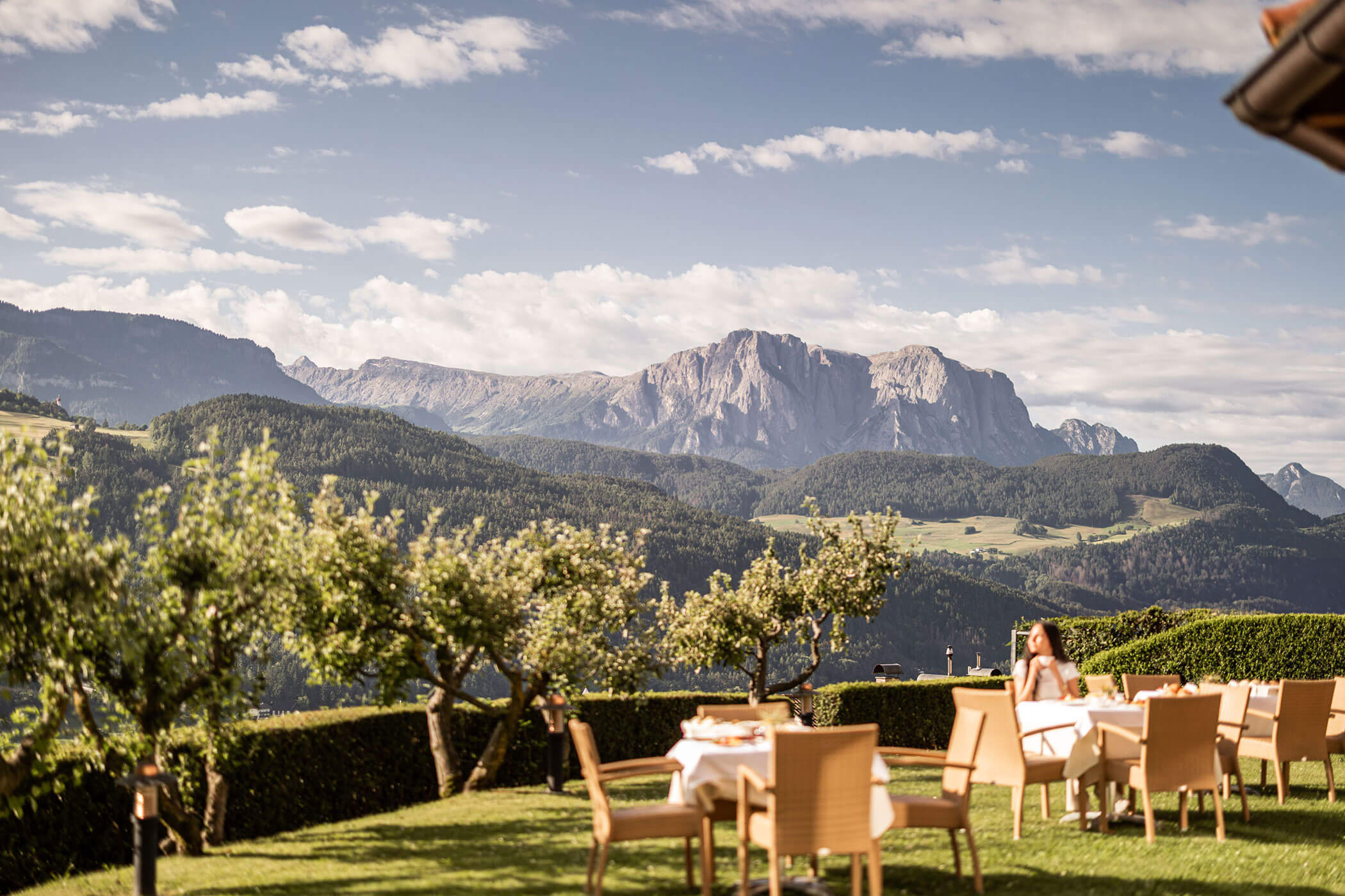 Essbereich im Freien mit Tischen und Stühlen mit Blick auf Berge und grüne Hügel unter einem teilweise bewölkten Himmel. - Egger Apartments & Suites