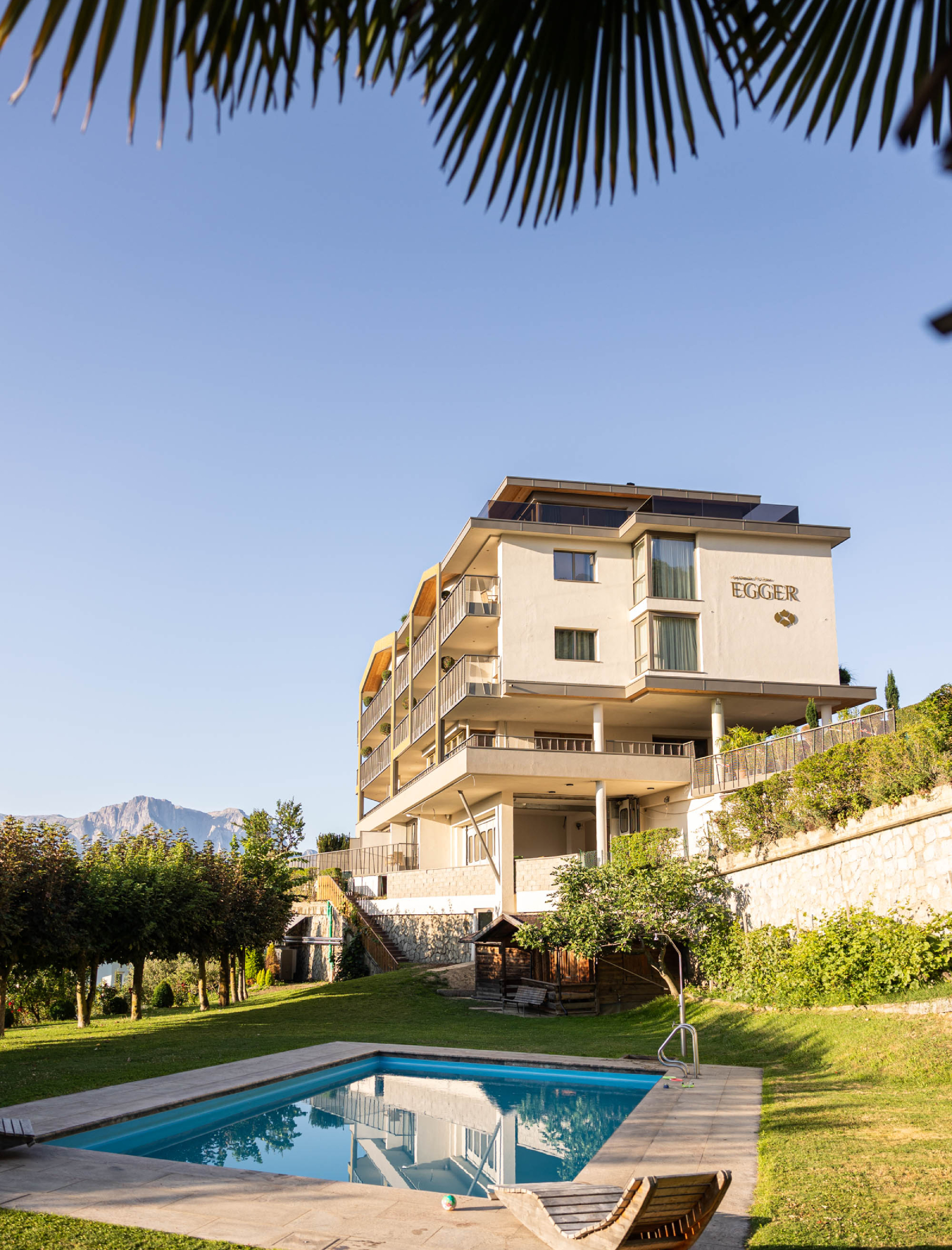 Modernes Apartmenthaus mit Balkonen in einem grünen, hügeligen Dorf mit einer Kirche und Bergen im Hintergrund. - Egger Apartments & Suites