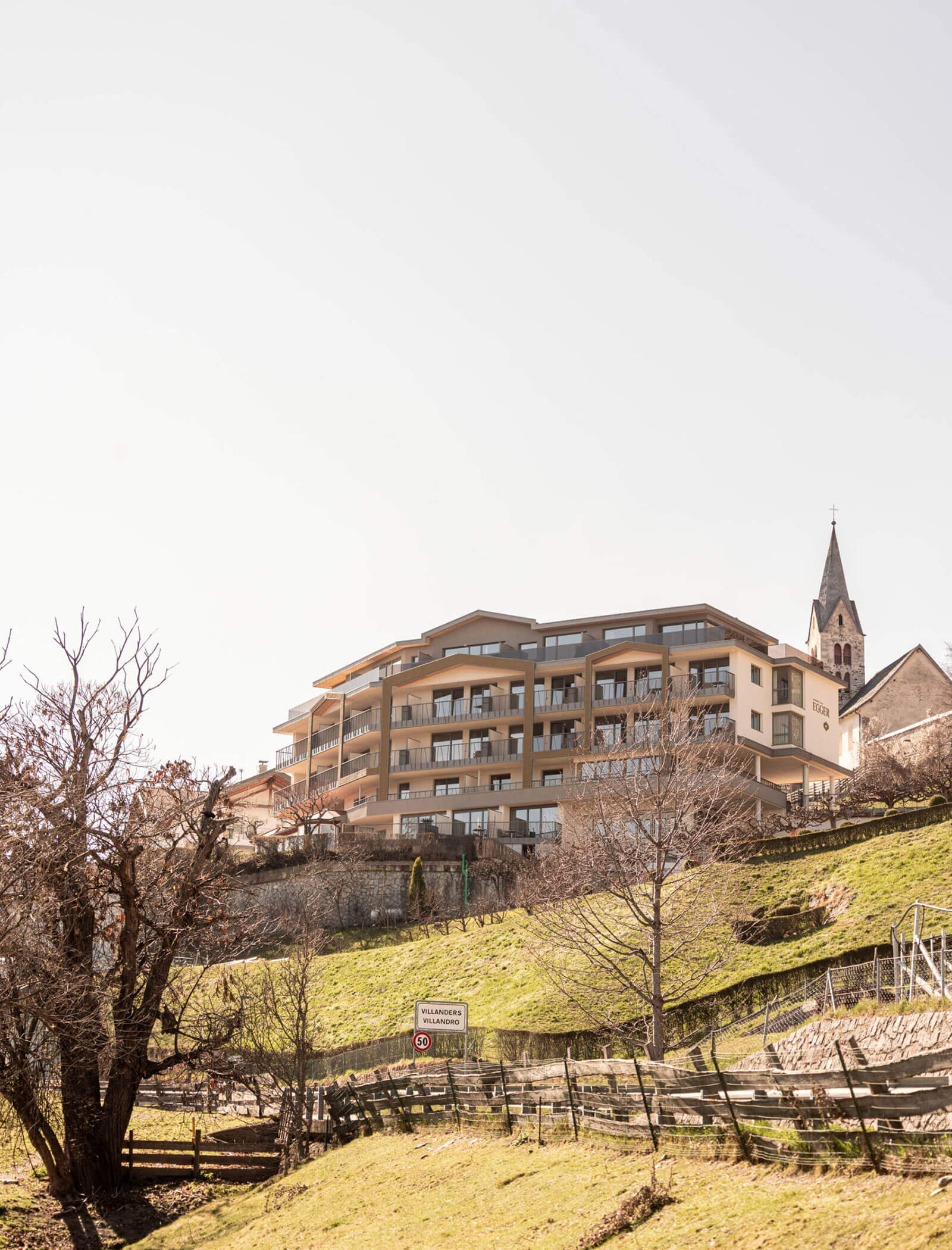 Modernes Apartmenthaus mit Balkonen in einem grünen, hügeligen Dorf mit einer Kirche und Bergen im Hintergrund. - Egger Apartments & Suites