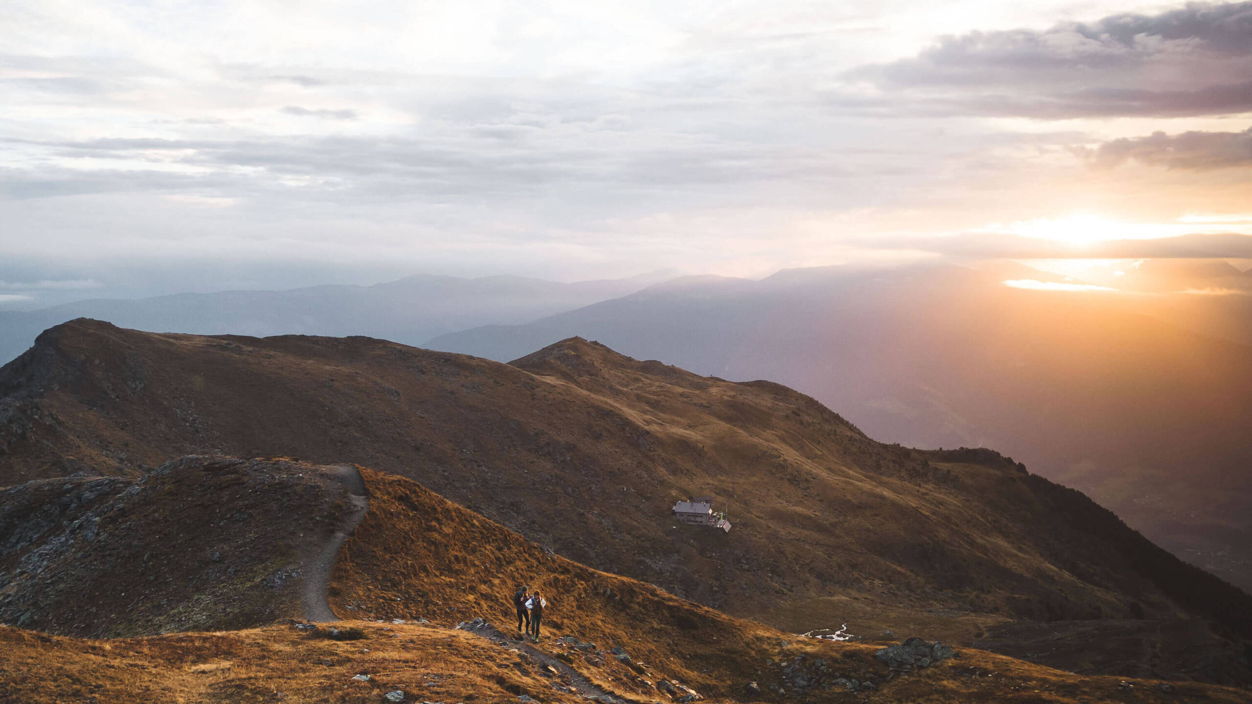 Zwei Wanderer stehen bei Sonnenuntergang auf einem Bergpfad und blicken auf sanfte Hügel und einen fernen, leuchtenden Horizont. - Egger Apartments & Suites