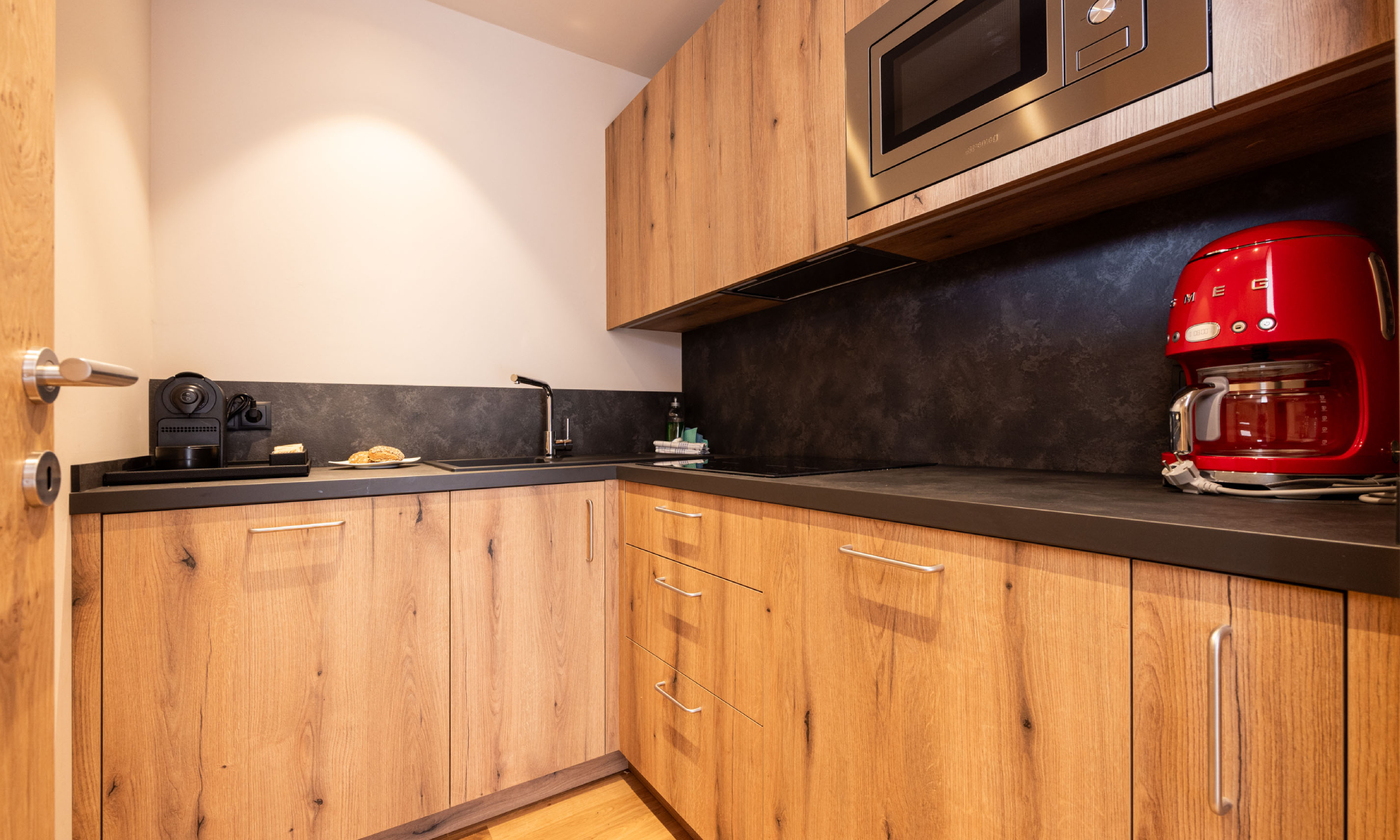 ausgestattete Küche aus Holz mit roter Kaffeemaschine, Espressomaschine, Mikrowelle, E-Herd, Spülmaschine, Geschirr und Tür zum schließen - Egger Apartments & Suites