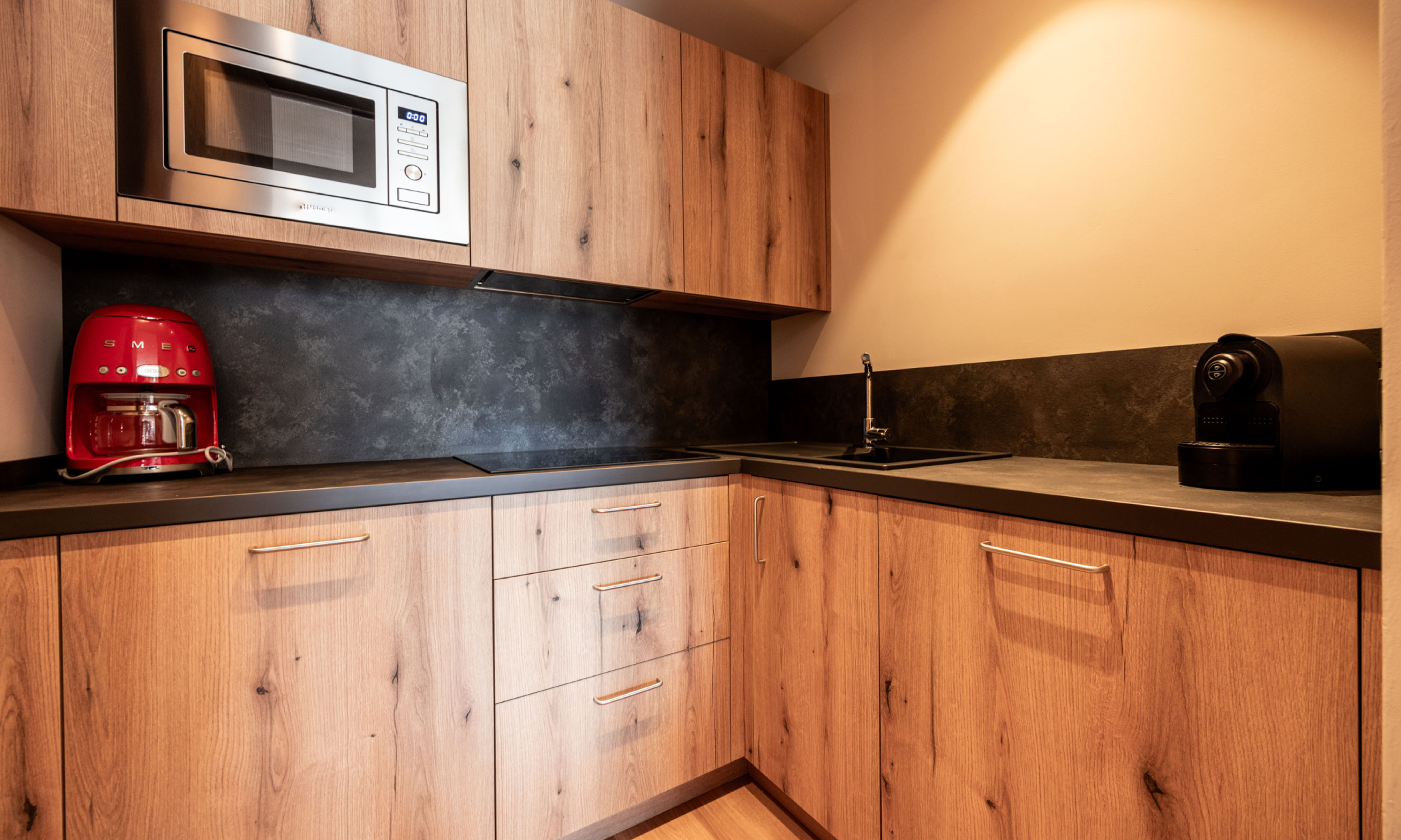 ausgestattete Küche aus Holz mit roter Kaffeemaschine, Espressomaschine, Mikrowelle, E-Herd, Spülmaschine, Geschirr und Tür zum schließen - Egger Apartments & Suites