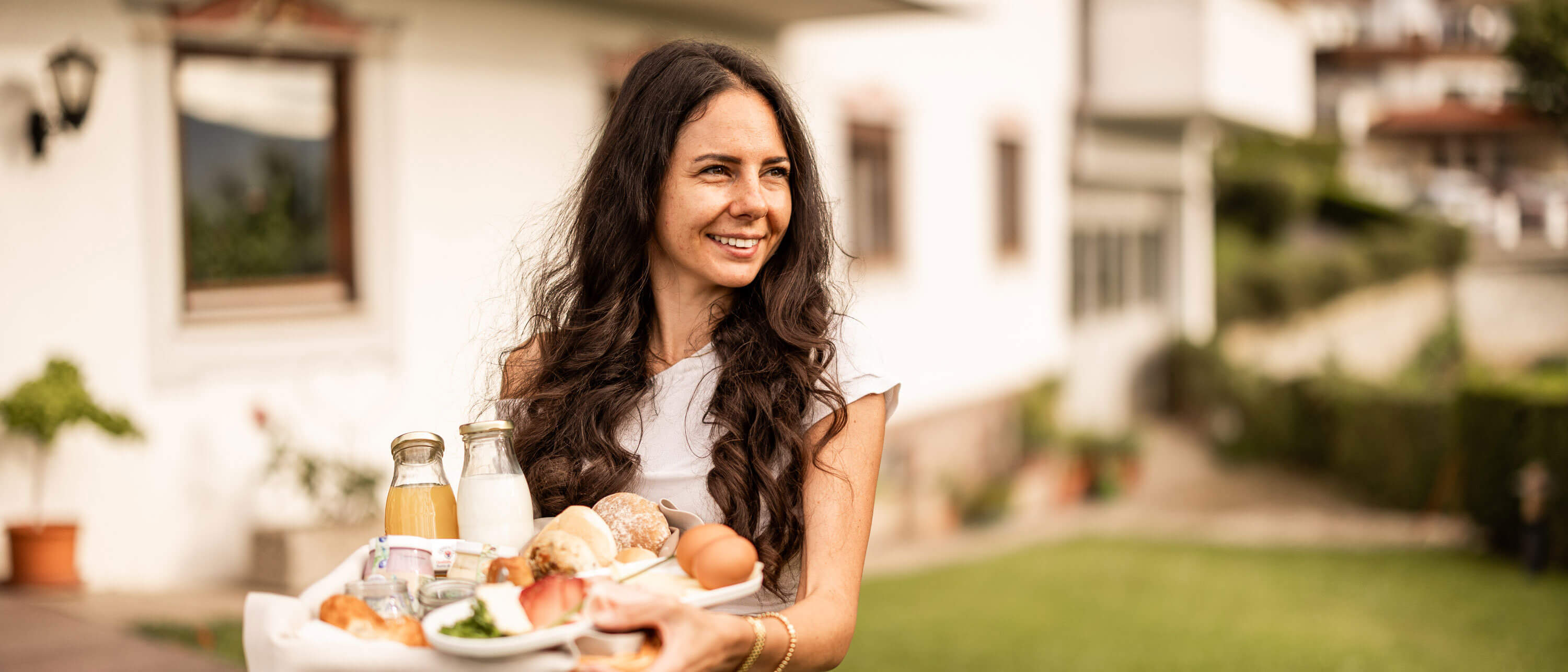 Eine Frau lächelt im Freien und hält ein Tablett mit Frühstück, Saft und Kaffee in einer Gartenanlage. - Egger Apartments & Suites