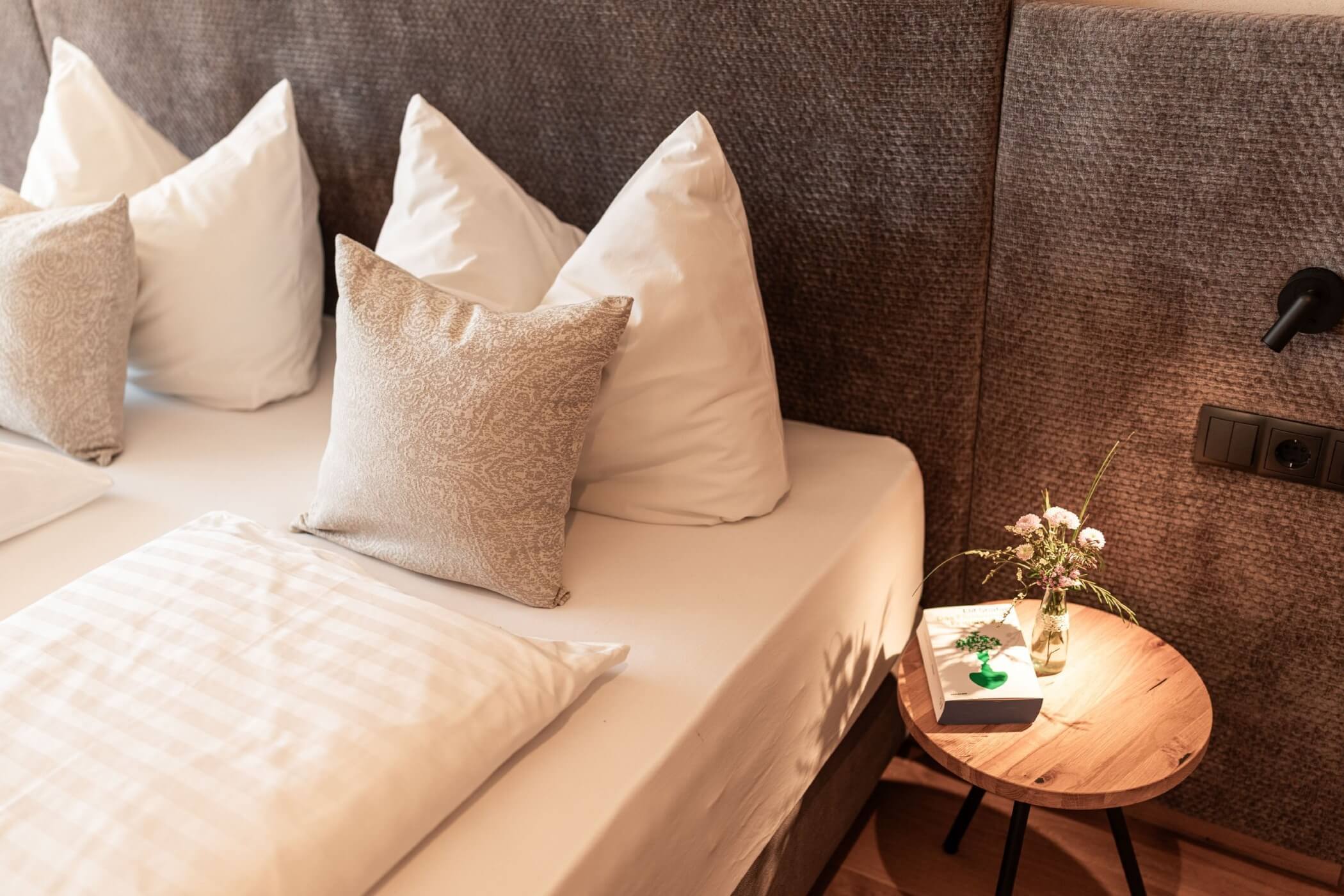 ein sauber gemachtes Bett mit einer Blumenvase und einem Buch auf dem Tisch daneben - Egger Apartments & Suites