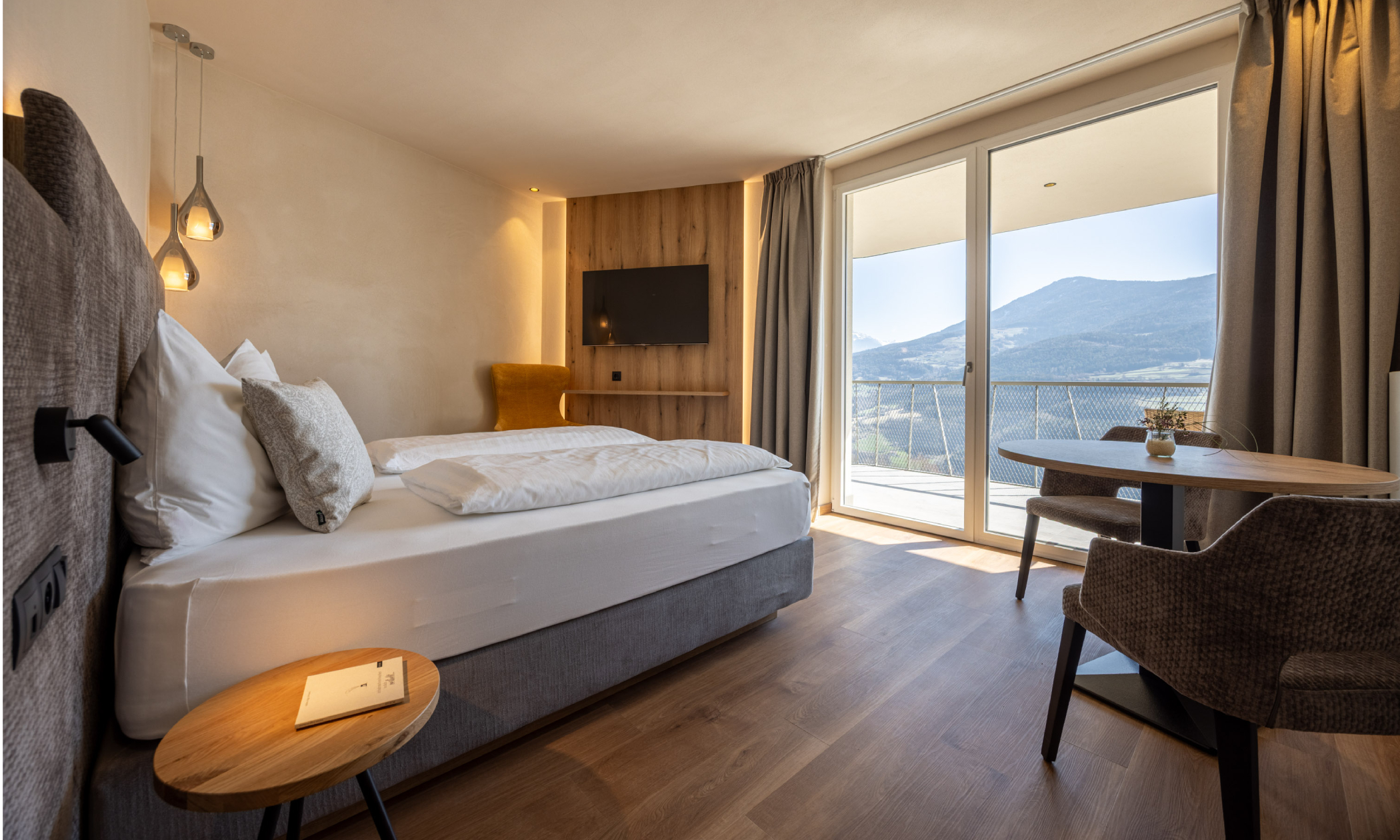 Modernes helles Schlafzimmer mit großen Fenstern und Balkon mit Bergblick, großem Bett, ockerfarbenen Sessel und Sitzecke - Egger Apartments & Suites